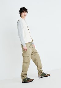 Vesta bej deschis peste un tricou gri cu mânecă lungă, asortată cu pantaloni cargo khaki și adidași verde-maro cu șireturi negre.