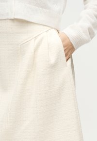 Χέρι στην τσέπη της υφασμάτινης κρεμ φούστας που φοριέται με ριμπ πλεκτό μανίκι κρεμ πουλόβερ ορατό σε ουδέτερο φόντο.