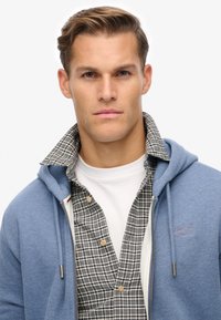 Haut à zip bleu superposé sur une chemise à carreaux gris et blanc, avec un t-shirt blanc à col rond en dessous. Il comporte un logo sur la poitrine.