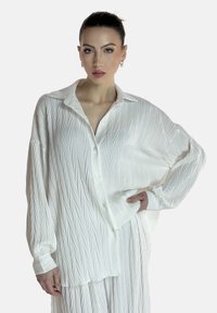 Chemise blanche à manches longues avec un design à boutons, présentant un motif texturé de lignes diagonales et une coupe décontractée, fabriquée en tissu léger.