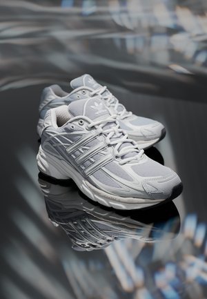 Pair of white Adidas athletic shoes with mesh and synthetic overlays resting on a reflective dark surface.
