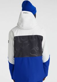 Blauwe en witte jas met een zwarte, gebloemde bovenkant, kap en manchetten. Bevat een logo op de achterste kraag. Glad, waterafstotend materiaal.