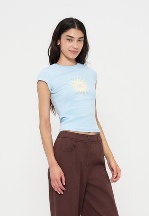 Ung kvinde iført en lyseblå tætsiddende t-shirt med en gul sol-grafik og løse brune bukser med høj talje, står mod en hvid baggrund.