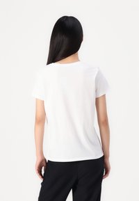 White cotton t-shirt, short sleeves, crew neck, relaxed fit, smooth texture, side slits, paired with black pants. Back view.