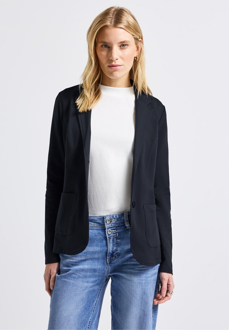 Černý blazer z lehkého materiálu, s klopovým límcem, dvěma předními kapsami a upraveným střihem, doplněný o bílé top s ribkovaným vzorem.