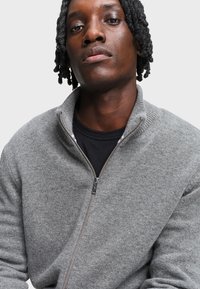 Jonge man met korte dreadlocks, gekleed in een grijze ritstrui over een zwart shirt, tegen een effen lichte achtergrond.