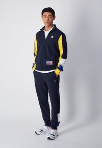 Tuta Champion in blu navy, giallo, bianco e blu; giacca con zip e logo; pantaloni abbinati; design sportivo; la texture del tessuto è liscia.