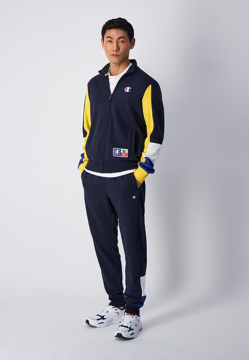 Tuta Champion in blu navy, giallo, bianco e blu; giacca con zip e logo; pantaloni abbinati; design sportivo; la texture del tessuto è liscia.