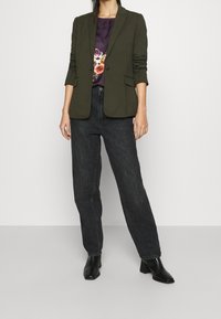 Mujer vestida con un blazer verde oliva sobre una blusa floral púrpura, jeans de tiro alto negros y botines negros de tacón, de pie contra un fondo blanco.