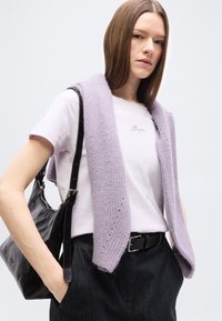 Νεαρή γυναίκα με ίσια μαλλιά, φοράει λευκό T-shirt, πλεκτό πουλόβερ λεβάντας ριγμένο στους ώμους, μαύρη ζώνη, σκούρο παντελόνι και μαύρη τσάντα ώμου.