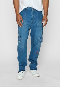 Blaue Denim-Cargo-Hosen mit lockerer Passform, ausgestattet mit mehreren Taschen, einem roten Logo-Patch und einer leicht abgewetzten Textur.