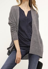 Cardigan gris de apertura frontal en un material de punto suave, con mangas largas y un ajuste relajado, superpuesto sobre una blusa azul marino y jeans de mezclilla.