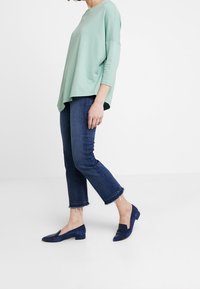 Maglione oversized verde chiaro e texturizzato con maniche a 3/4, abbinato a jeans cropped blu scuro con orli sfrangiati e scarpe basse navy.