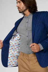 Giacca blu a righe con fodera interna a motivo, indossata su una camicia grigia con bottoni. I pantaloni beige completano l'outfit.
