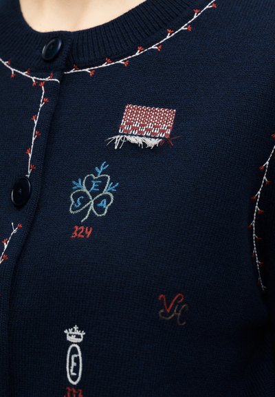 Μπλε ναυτικό πλεκτό ζακέτα με μικρά κεντήματα που περιλαμβάνουν ένα μπαλώμα, τριφύλλι με γράμματα και διακοσμητικές ραφές κοντά σε μαύρα κουμπιά.