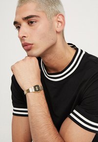 Goldene Digitaluhr mit quadratischem Zifferblatt und Metallarmband, getragen an einer Hand, die gegen ein schwarzes Hemd mit weißen Streifenakzenten ruht.