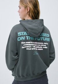 Sudadera gris con capucha ajustable. Presenta un gran texto en color teal y blanco en la parte posterior con frases motivacionales y un detalle de firma.