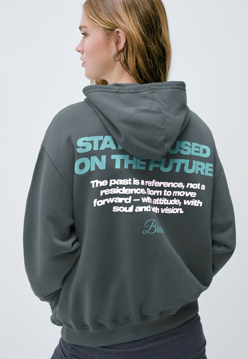 Sudadera gris con capucha ajustable. Presenta un gran texto en color teal y blanco en la parte posterior con frases motivacionales y un detalle de firma.
