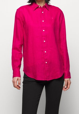 Person trägt ein hellpinkes, langärmliges Hemd mit Knopfleiste und Kragen sowie eine schwarze Hose, steht vor einem einfarbigen Hintergrund.