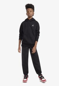 Black hoodie with a front pocket and white logo, paired with black sweatpants and red and white sneakers. Short, curly hairstyle.