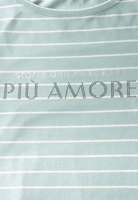 Pruhované světle modré tričko s bílými vodorovnými pruhy. Obsahuje text "storie delle vacanze" ve stříbrné barvě a "PIÙ AMORE" z kamínků.