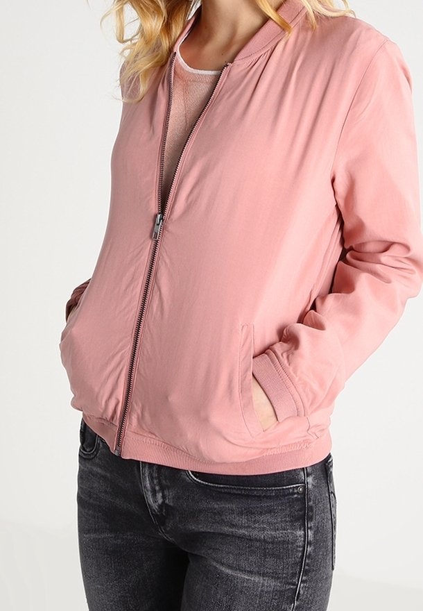 Jasnoróżowa kurtka bomberka z zapięciem na zamek z przodu, bocznymi kieszeniami, ściągaczami przy mankietach i gładką teksturą, noszona na lekką bluzkę i ciemne dżinsy.