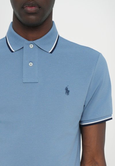Vaaleansininen golfpaita, joka on valmistettu teksturoidusta puuvillasta. Paita sisältää kolmen napin kaula-aukon ja kontrastoivan laivastonsinisen ja valkoisen kauluksen. Rinnassa brodeerattu logo.