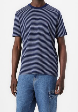Mand iført navyblå og hvidstribet kortærmet T-shirt og blå denim cargobukser, står med armene afslappet ned langs siderne.
