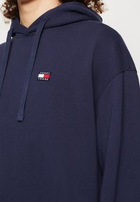 Navy blue hoodie made of cotton blend, featuring a logo patch in red, white, and blue. It has a drawstring hood and a smooth texture.