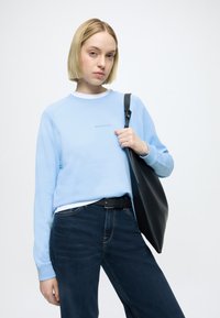 Šviesiai mėlynas džemperis su apvalia iškirpte, su ribotais rankogaliais ir siuvinėta logotipu, derinamas su tamsiai mėlynais džinsais ir juoda pečių krepšiu.