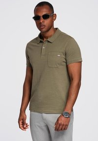 Polo shirt verde oliva con colletto, patta con bottoni e una singola tasca sul petto. Il modello indossa occhiali da sole scuri e un orologio argento.
