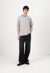 T-shirt a righe a maniche corte in grigio chiaro e grigio scuro, abbinato a jeans neri a gamba larga. Presenta un piccolo dettaglio del logo nero sulla maglietta.