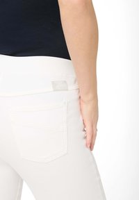 Witte jeans met een gladde textuur, een brede rekbare tailleband en twee achterzakken. Kenmerken zijn een klein logo label op de tailleband.
