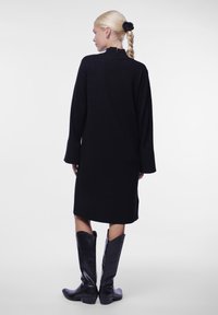 Juoda ilgomis rankovėmis suknelė su aukštu kaklu ir šonine įpjova, derinama su kelių aukščio juodais odiniais batais, turinčiais smailų dizainą ir lenktas kraštus.
