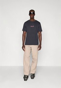 Melna īsroku krekls ar sarkanu ķiršu grafiku, kopā ar gaiši bēšām kareivju biksēm un melniem apaviem, modelis valkā saulesbrilles.