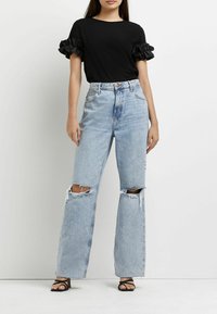 Camiseta negra con mangas rizadas, combinada con jeans de pierna ancha, rotos y de color azul claro; unas sandalias negras de tacón abierto completan el atuendo.