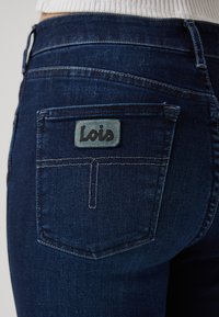 Tamsiai mėlyni džinsai su galine kišene, turinčia kvadratinę etikėtę su užrašu "Lois" juodu tekstu. Detalizuotos siūlės ir glotni tekstūra.
