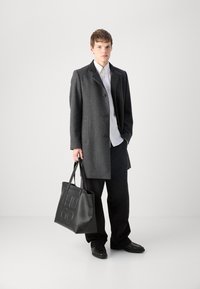 Grauer Wollmantel mit aufgeschlagenem Kragen und schwarzem Knopfverschluss, kombiniert mit einem weißen Hemd, schwarzen Hosen und einer schwarzen Ledertasche.