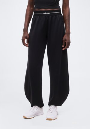 Osoba nosící černé široké kalhoty Adidas s elastickým pasem a bílé tenisky, stojící na jednobarevném pozadí.