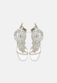 Sandales argentées à lanières avec des ailes en strass décoratives, des brides fines et des semelles rembourrées. Elles présentent une finition métallique lisse et des détails élaborés.