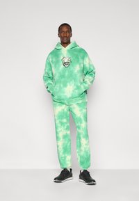 Πράσινο φούτερ με γραφικό τύπωμα και αντίστοιχο παντελόνι φόρμας. Το υλικό φαίνεται απαλό, με μείγμα ανοιχτόχρωμων και σκούρων πράσινων τόνων.