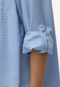 Lichtblauw, gestructureerd shirt met opgerolde mouwen die met knopen zijn vastgezet. Close-up met detail van de stof en arm.
