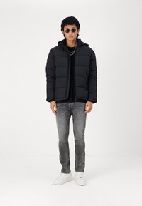 Svart pufferjakke med hette, med et kviltet design. Brukt over en svart skjorte, kombinert med grå jeans og hvite sportssko.