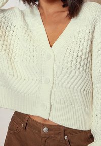 Vit stickad cardigan med V-ringning, tre knappar, strukturerade vertikala och zick-zack mönster samt ribbad nederkant. Bärs över bruna byxor.