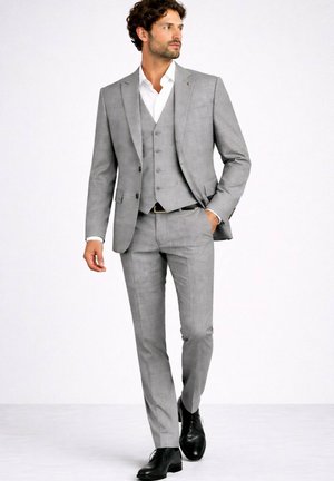 Homme marchant portant un costume trois pièces gris clair avec une chemise blanche et des chaussures habillées noires sur un fond blanc uni.