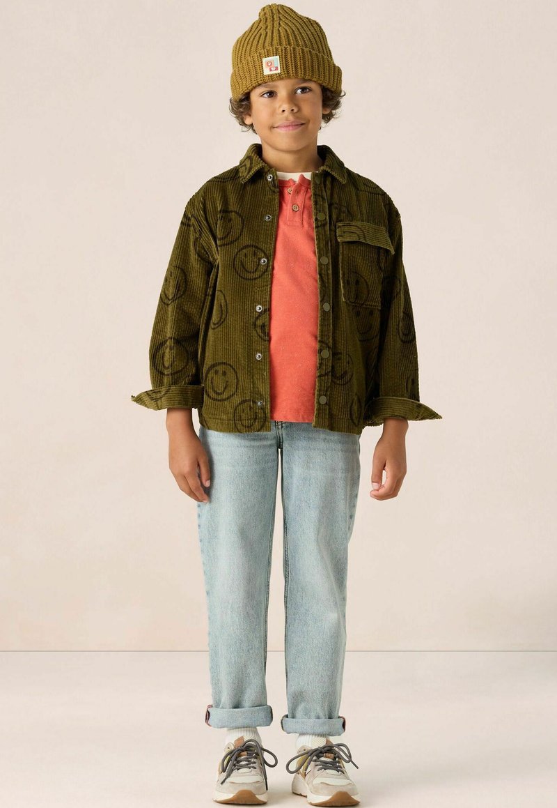 Kind dat een groen corduroy shirt met een glimlachend gezichtpatroon draagt, een oranje t-shirt, lichtblauwe opgerolde jeans en beige sneakers met grijze accenten.