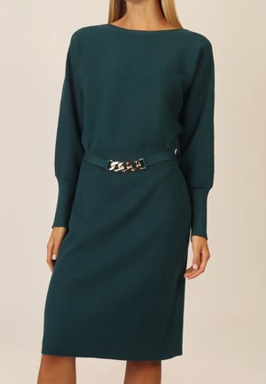 Femme portant une robe en maille côtelée couleur sarcelle à manches longues avec une ceinture chaîne dorée à la taille, posant devant un fond uni.