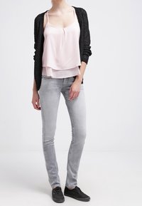 Gilet en tricot noir par-dessus une camisole à plusieurs couches rose clair. Modèle portant un jean gris ajusté et des sneakers noirs. Textures lisses avec un contraste de couleurs douces.