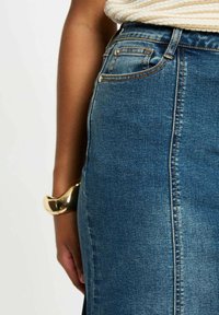 Gonna di jeans di un blu medio con una vestibilità dritta, caratterizzata da uno spacco laterale, dettagli cuciti e design a quattro tasche. Bracciale dorato visibile.