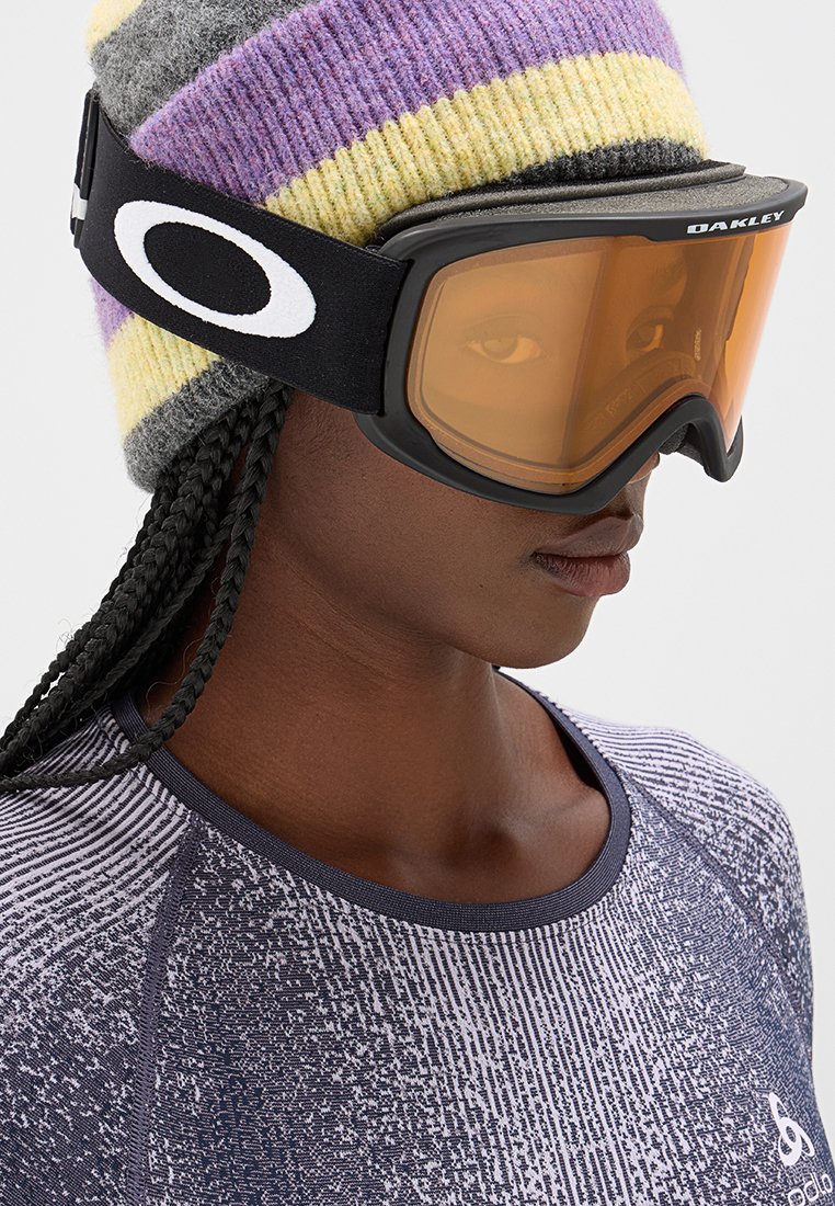 Osoba nosząca pasiasty fioletowy, żółty i szary czapkę-beanie, pomarańczowe gogle narciarskie Oakley oraz wzorzystą szaro-fioletową koszulę.